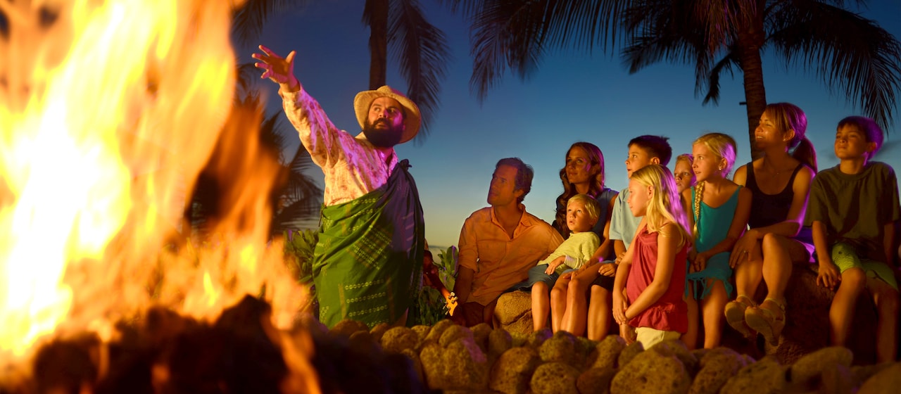 A man surrounded by palm trees performs a ceremony near a bonfire at sunset