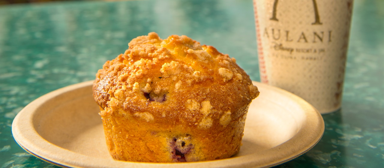 A blueberry muffin with a crumb topping on a plate beside a cup bearing the Aulani logo