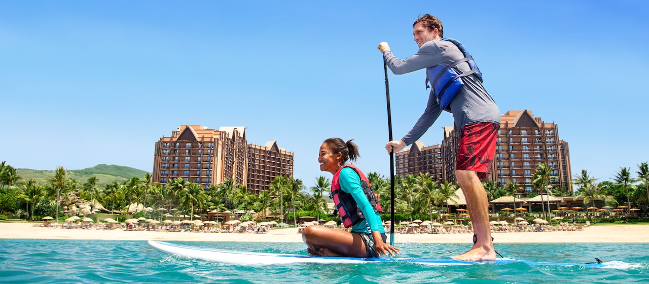 In the lagoon at Aulani Resort, a man stands and paddles a paddleboard as a woman rider sits on hers