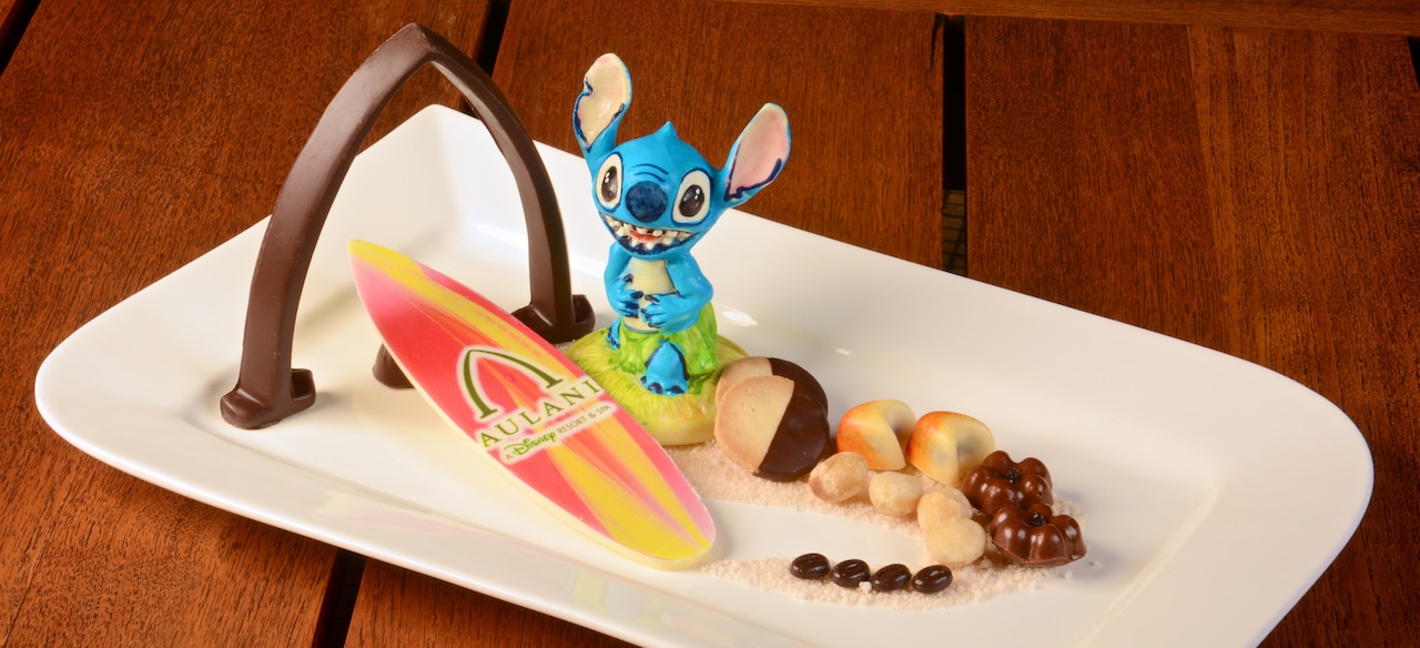 A plate of small treats is adorned with a chocolate arch, a replica surfboard and a Stich figurine.