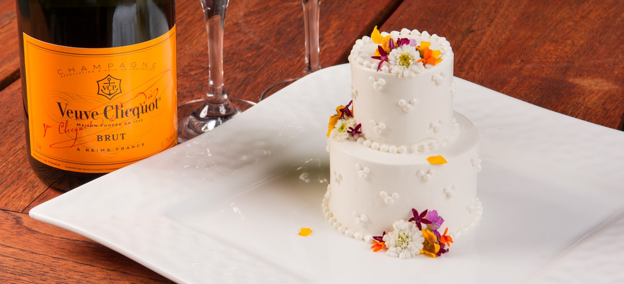 An elegantly decorated 2-tier iced mini cake served on a platter beside a bottle of Champagne.