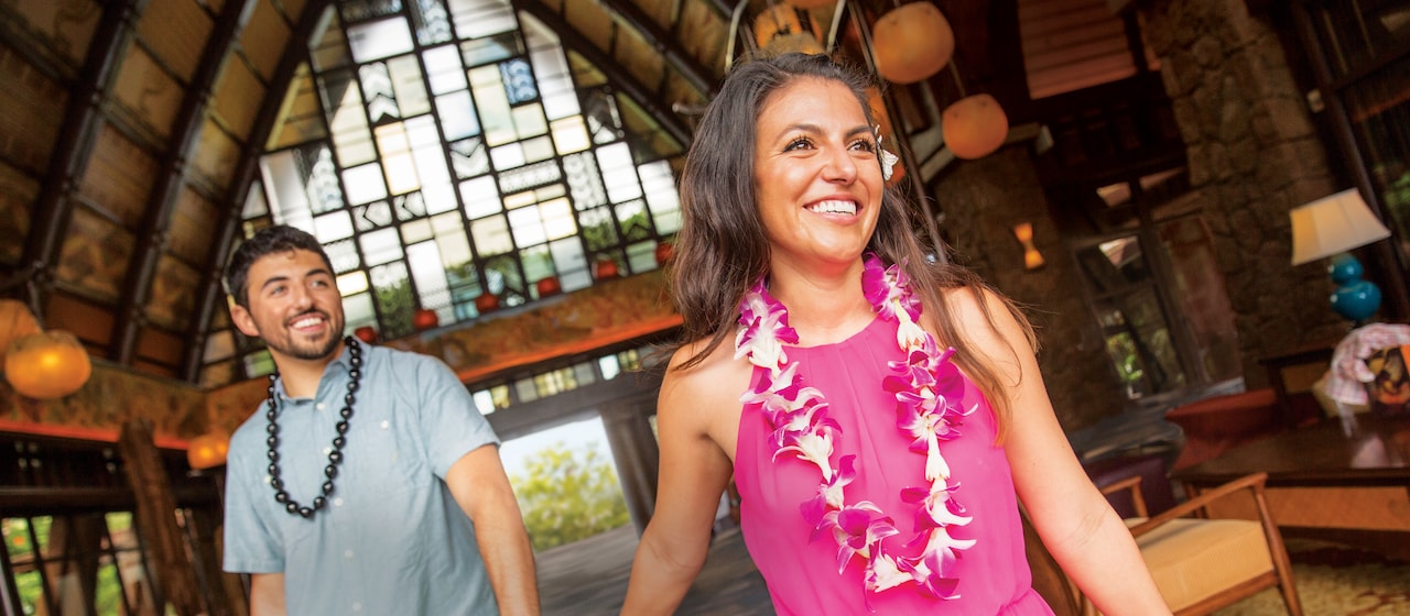 A woman wearing a Hawaiian flower lei holds hands with a man with a Kukui nut lei as they walk through the lobby of Aulani Resort