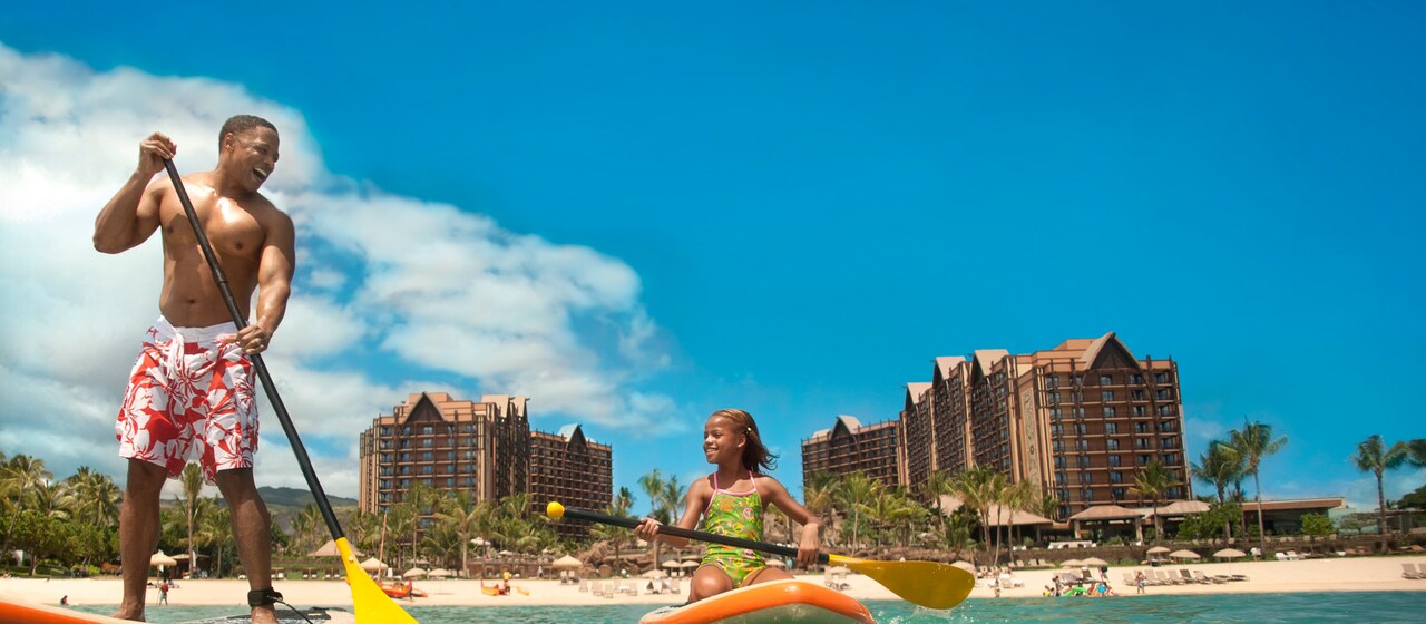 A father stands on a paddleboard next to his daughter, who is kneeling on a paddleboard, on the ocean in front of Aulani Resort