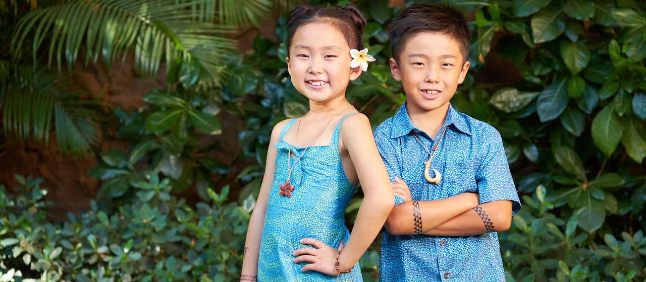 A young boy with tattoos on his crossed arms and a fish hook necklace stands next to girl with a flower behind her ear