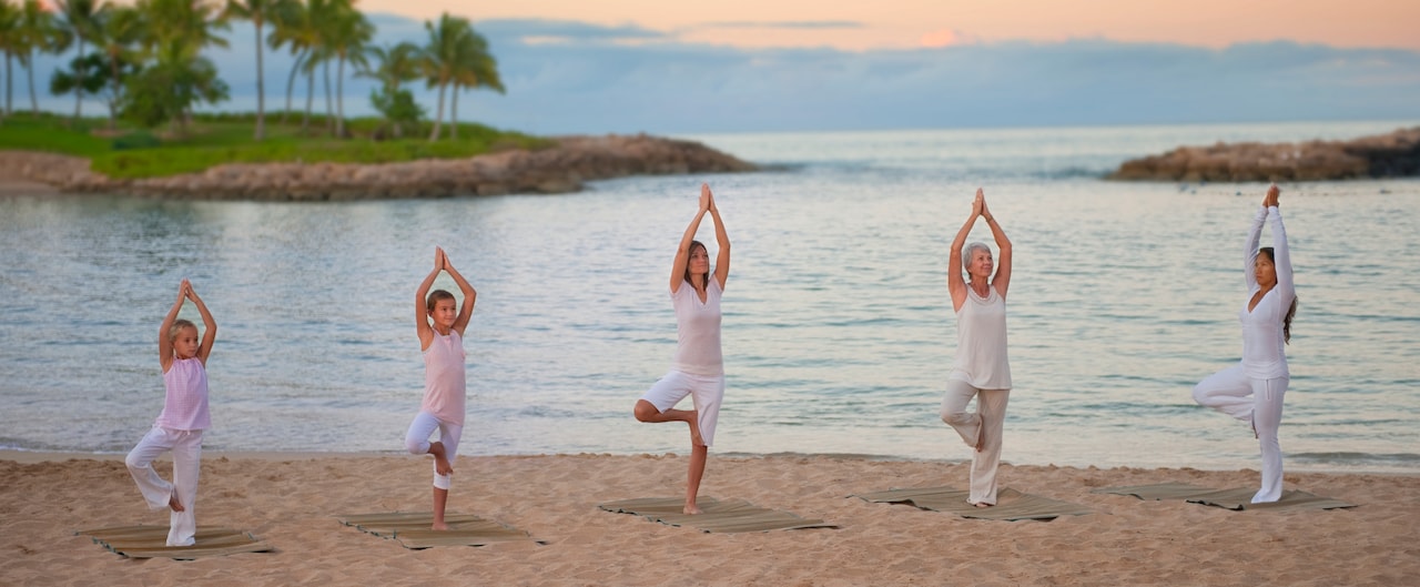 Two young girls and 3 women of various ages stand in tree pose in a yoga class on the beach