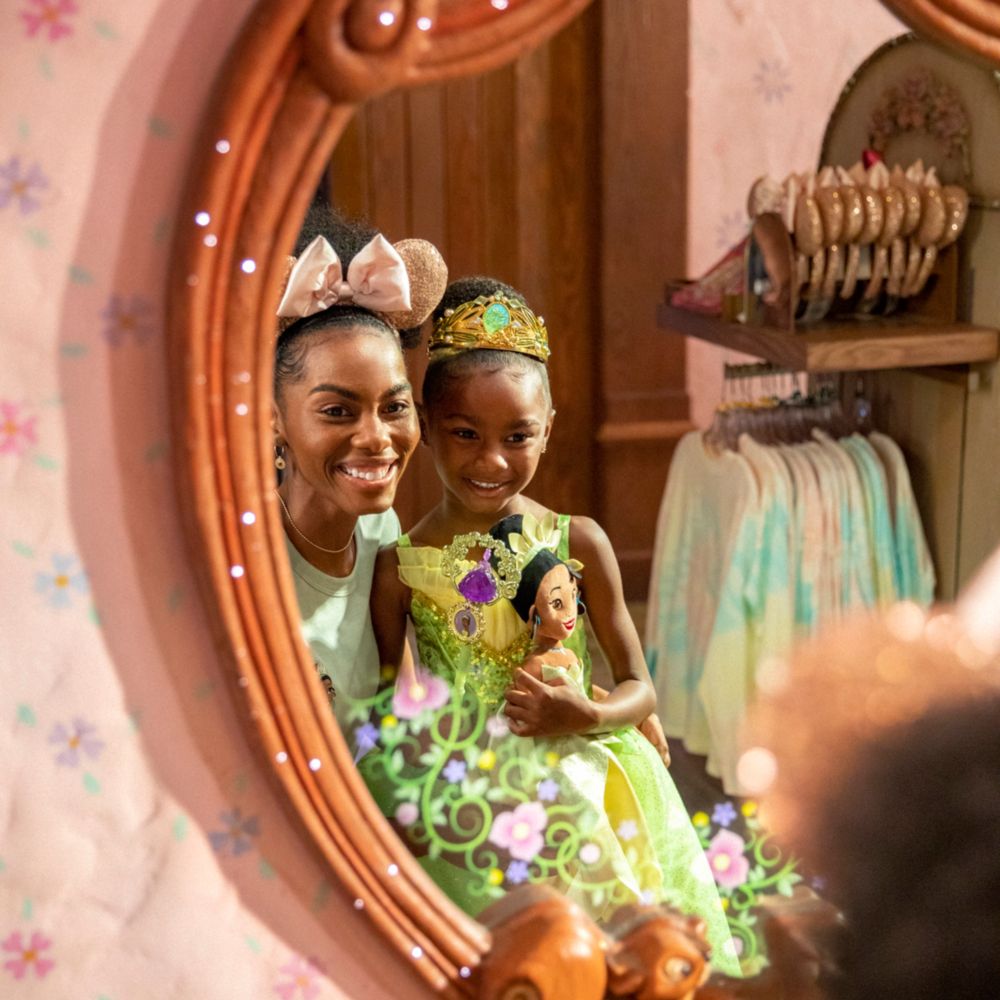 A mother looks in the mirror with her daughter who is dressed like Princess Tiana and holding a Princess Tiana plush doll 