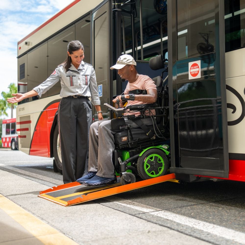 Un Miembro del Elenco guía a un hombre que usa una silla de ruedas motorizada al salir de un autobús del hotel usando una rampa.
