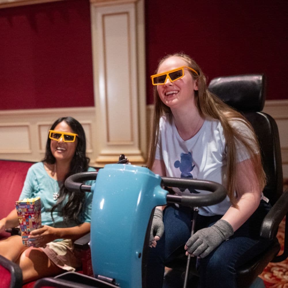A woman on a scooter sits beside a woman in a theatre seat, both wearing 3D glasses and smiling.