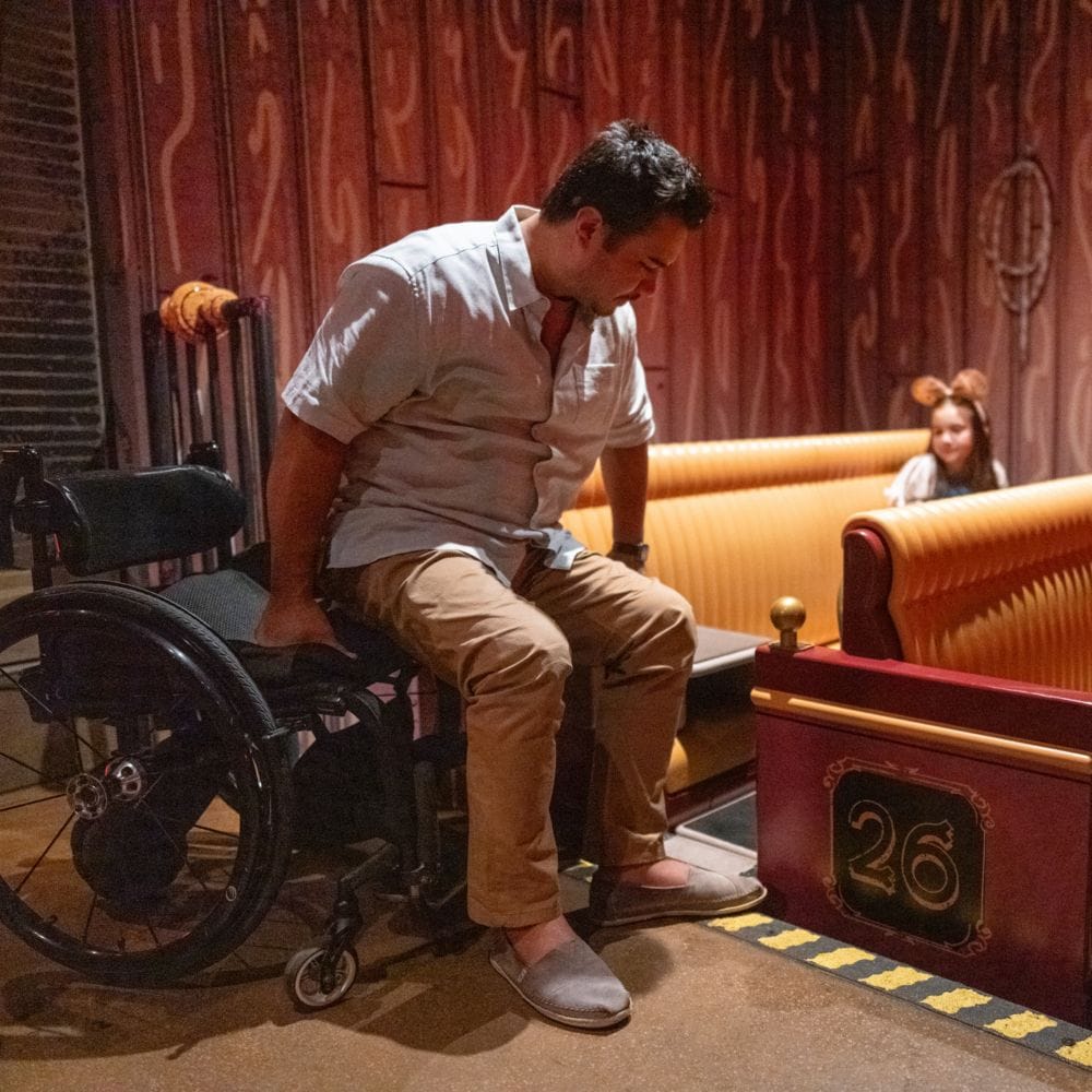 A man is sliding himself from his wheelchair onto the vehicle seat for Mickey and Minnie's Runaway Railway while a younger girl looks on from the vehicle.