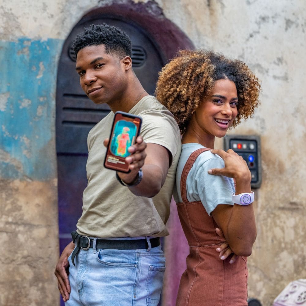 A woman wearing a Magic Band Plus standing back to back with a man holding out a phone