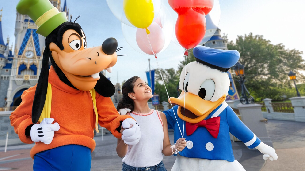 A young Guest holding 3 balloons walks between Goofy and Donald Duck in front of Cinderella Castle