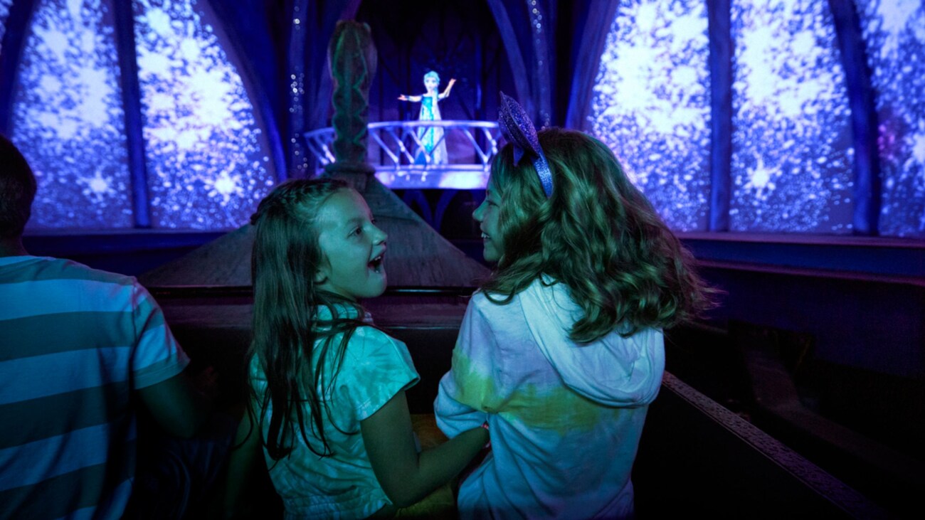 2 young girls look at one another with excitement while they ride the Frozen Ever After attraction as a figure of Elsa sings