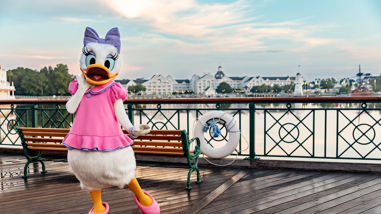 Daisy Duck posant dans une robe et un nœud sur une promenade au bord de l’eau avec bancs et Disney’s Yacht Club Resort et Disney’s Beach Club Resort au loin