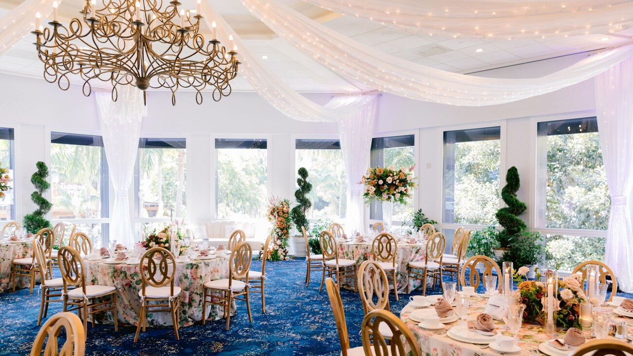 A ballroom event venue with a chandelier, dining tables, floral arrangements, topiaries, place settings and large windows looking out at a garden