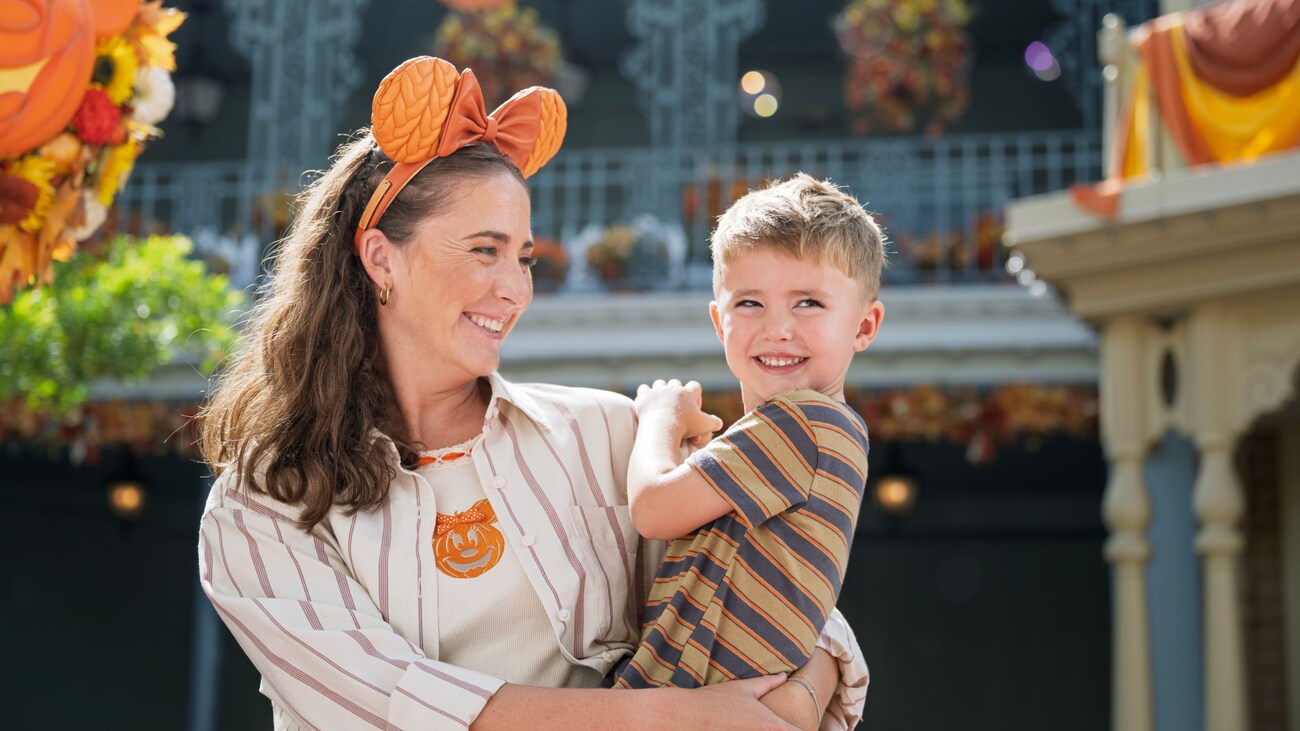 A Guest wearing a Minnie Mouse ear headband while holding a young boy on Main Street USA decorated with fall foliage