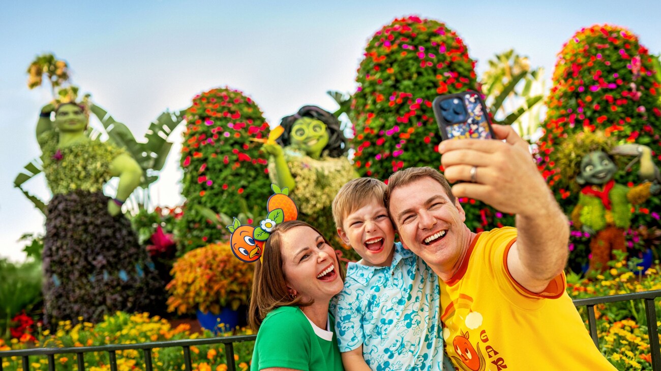Una familia de 3 integrantes se toma una selfi frente a los arbustos artísticos de los personajes de Encanto en el EPCOT International Flower & Garden Festival