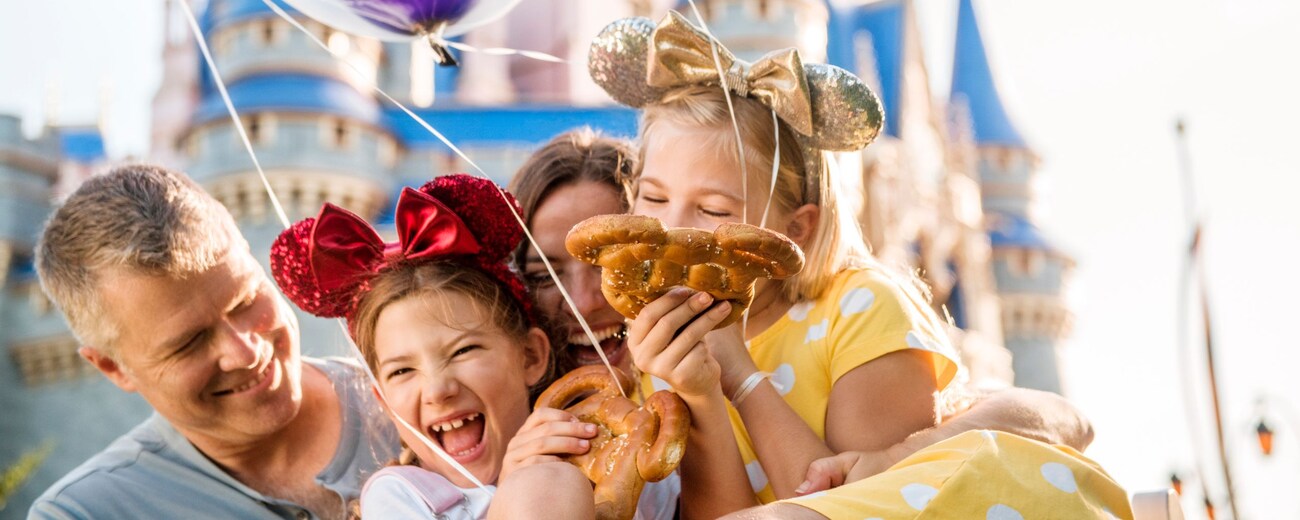 A family smiling and embracing each other while holding balloons and Mickey Mouse pretzels in front of Cinderella Castle