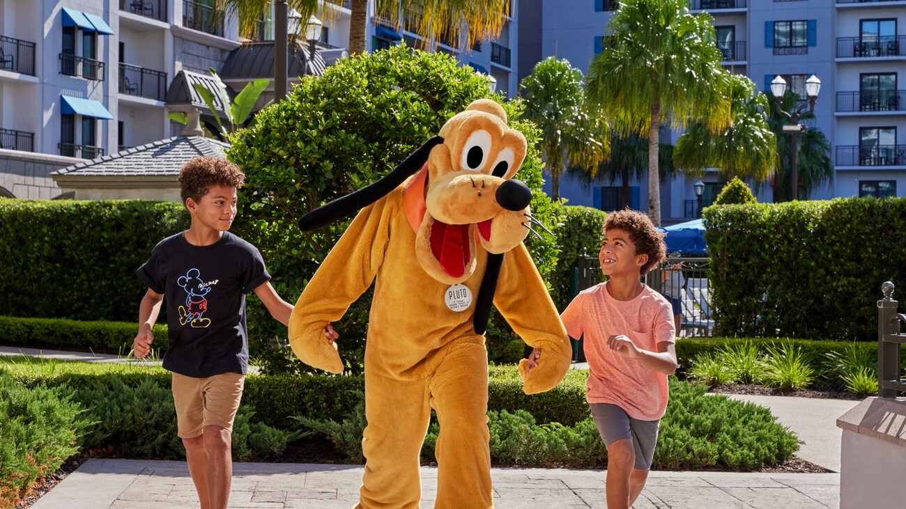 2 young boys walking hand in hand with Pluto along a paved path at Disney’s Riviera Resort