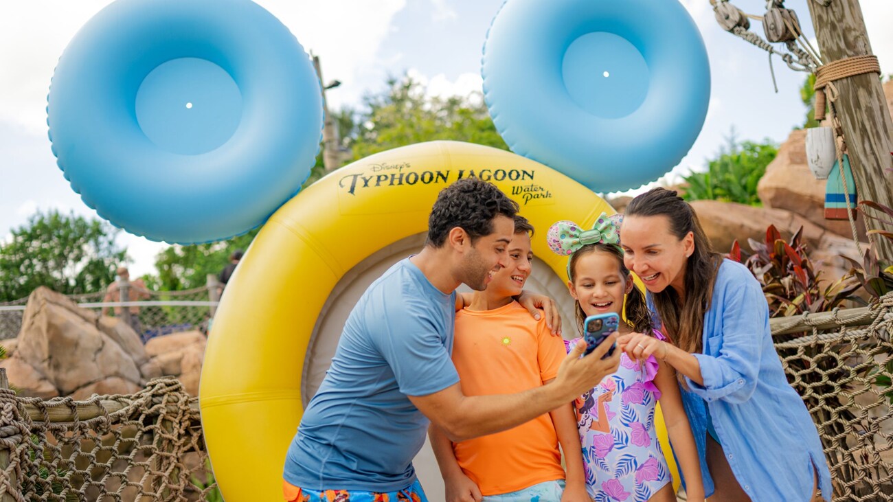 A family of 4 looking at a mobile device while standing next to an inner tube at Disney’s Typhoon Lagoon water park