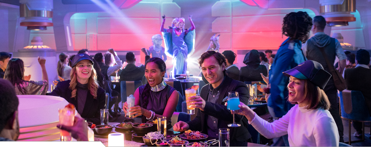 Guests raise a toast at a table while an intergalactic entertainer performs on stage in the background aboard the Halcyon starcruiser