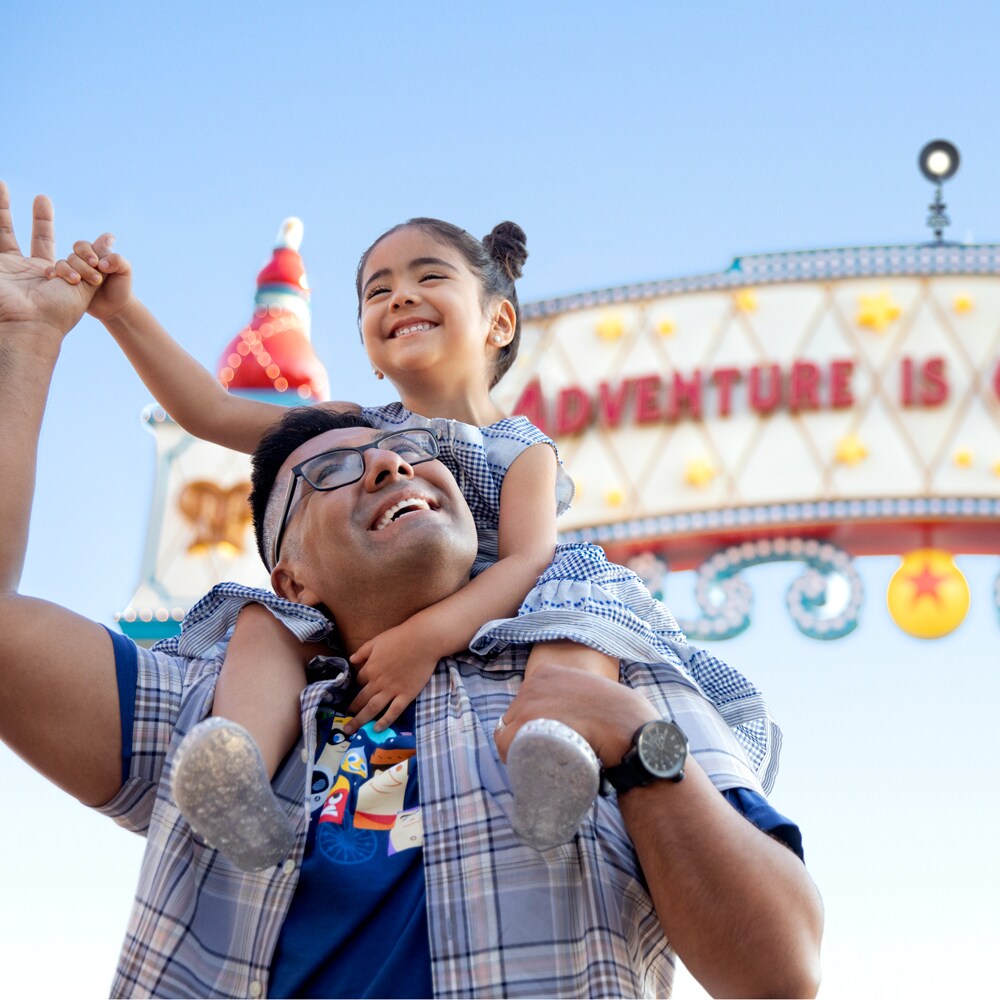 A smiling girl holds her father's hand while on his shoulders in front of a sign that reads