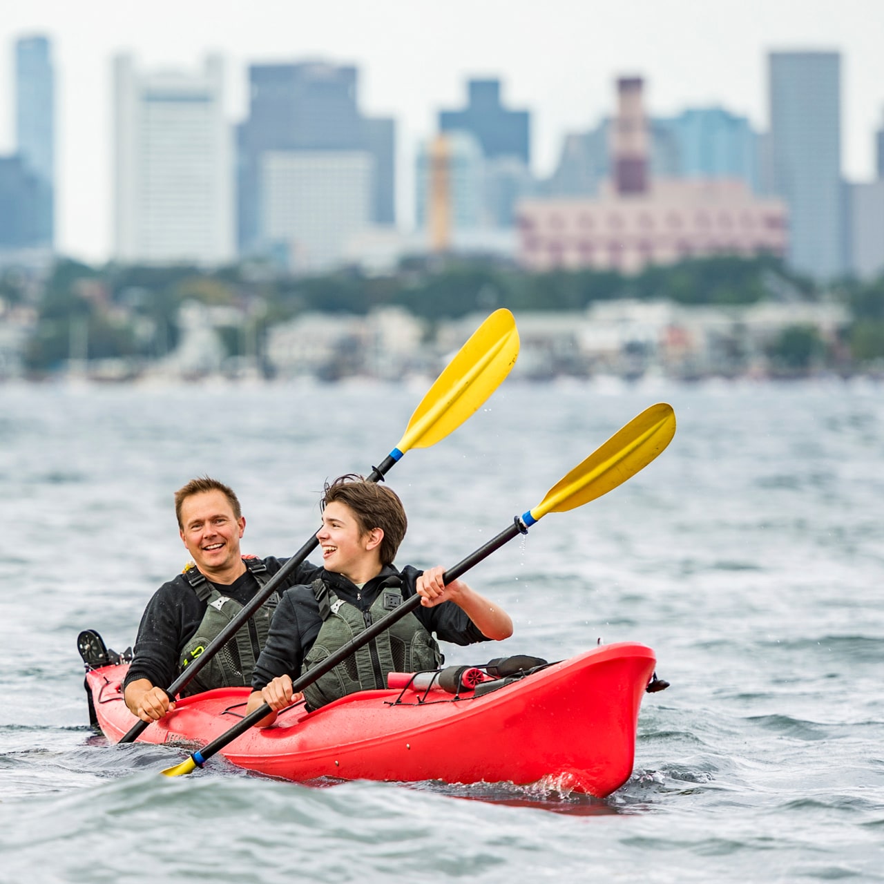 Two Adventurers in kayaks row in the water with the Boston skyline in the background
