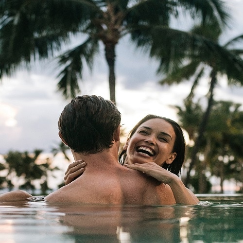 Two smiling adults in an infinity pool, with palms silhouetted along the shoreline