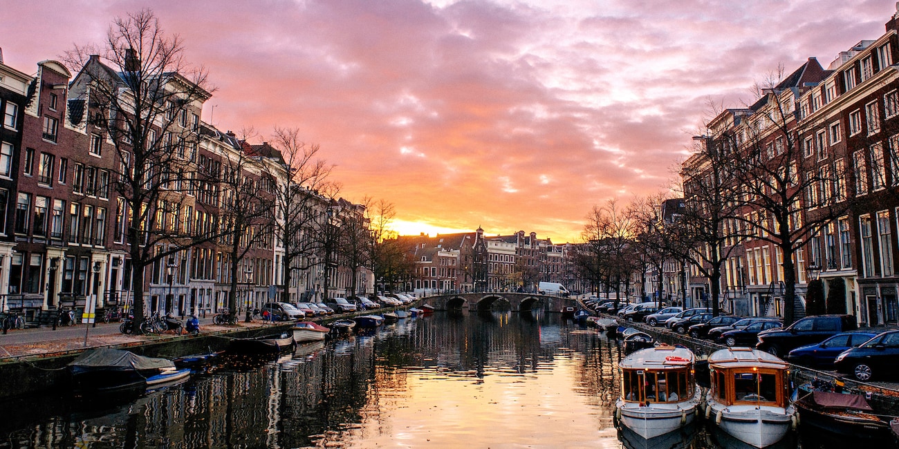A canal with parked boats runs through 2 building-lined streets in Amsterdam