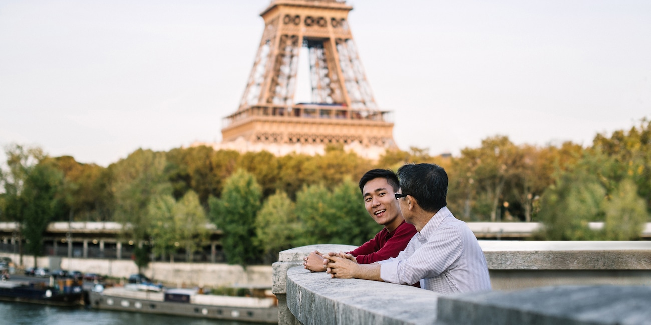 Two men stand with their arms resting on a cement railing overlooking the Seine River with the Eiffel Tower in the background