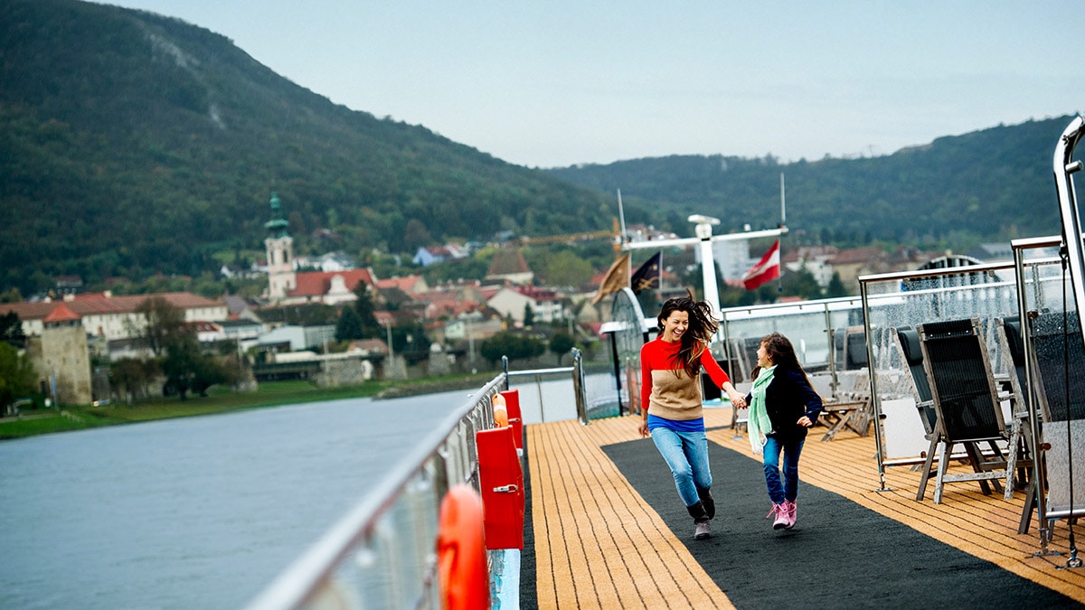 A mom and her young daughter laugh as they walk down the upper deck of a river cruise ship with a backdrop of a town between the riverbank and tree lined hills