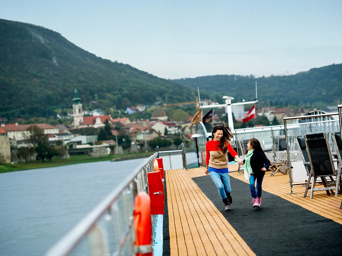 A mom and her young daughter laugh as they walk down the upper deck of a river cruise ship with a backdrop of a town between the riverbank and tree lined hills