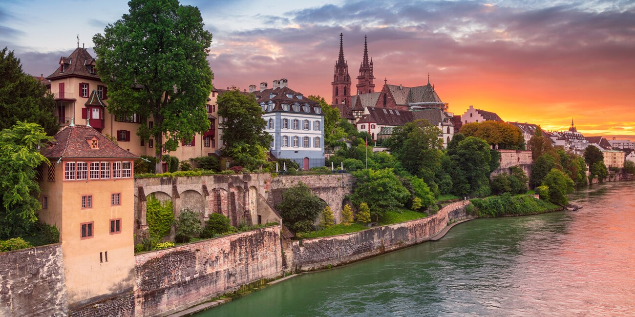 Two church spires stand above the buildings and houses of Basel, Switzerland along the banks of the Rhine River at sunset