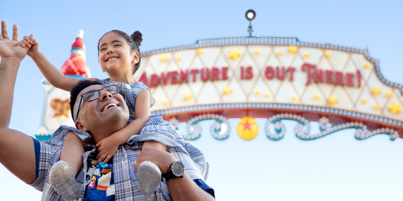 A smiling girl holds her father's hand while on his shoulders in front of a sign that reads