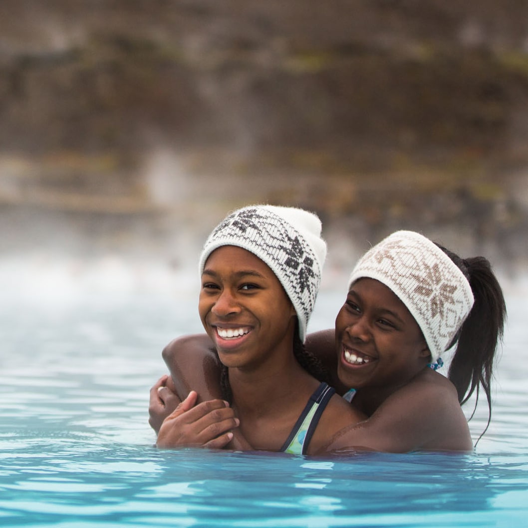 Two girls in matching headbands wade in an outdoor bath of steaming water near a man and a woman with a mountain in the background
