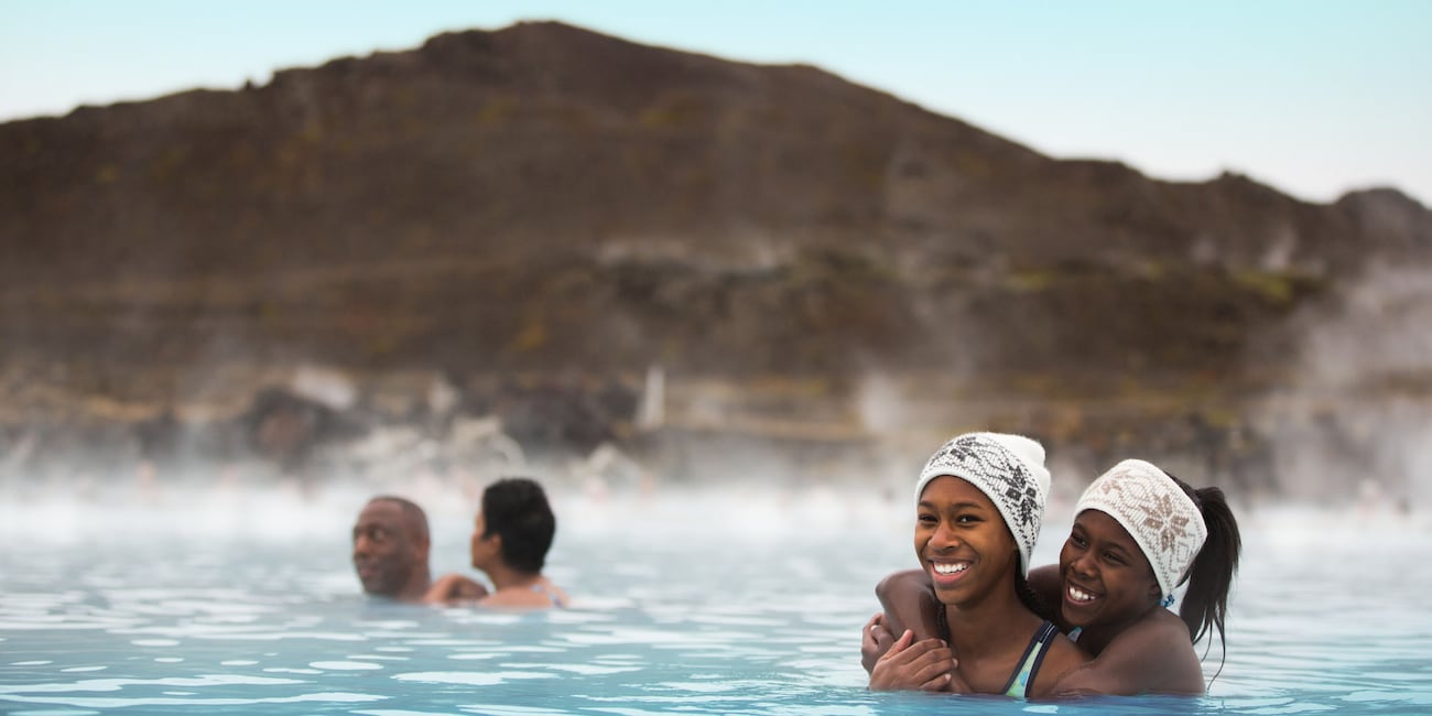 Two girls in matching headbands wade in an outdoor bath of steaming water near a man and a woman with a mountain in the background