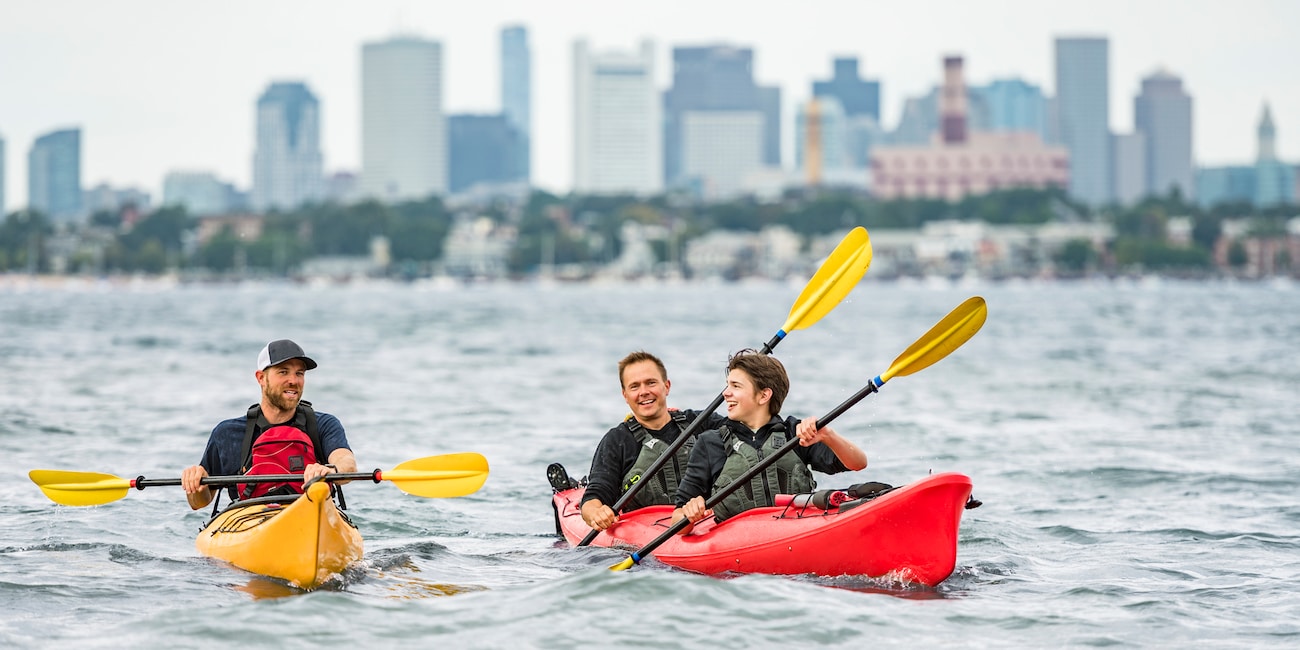 Two Adventurers in kayaks row in the water with the Boston skyline in the background