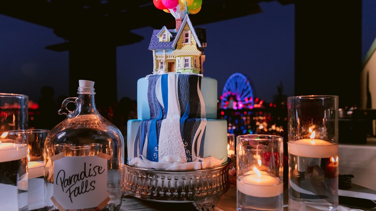 Una mesa con velas flotantes, una jarra de vidrio con una etiqueta que dice “Paradise Falls” y un pastel decorado con una cascada y la casa voladora de la película “Up” 