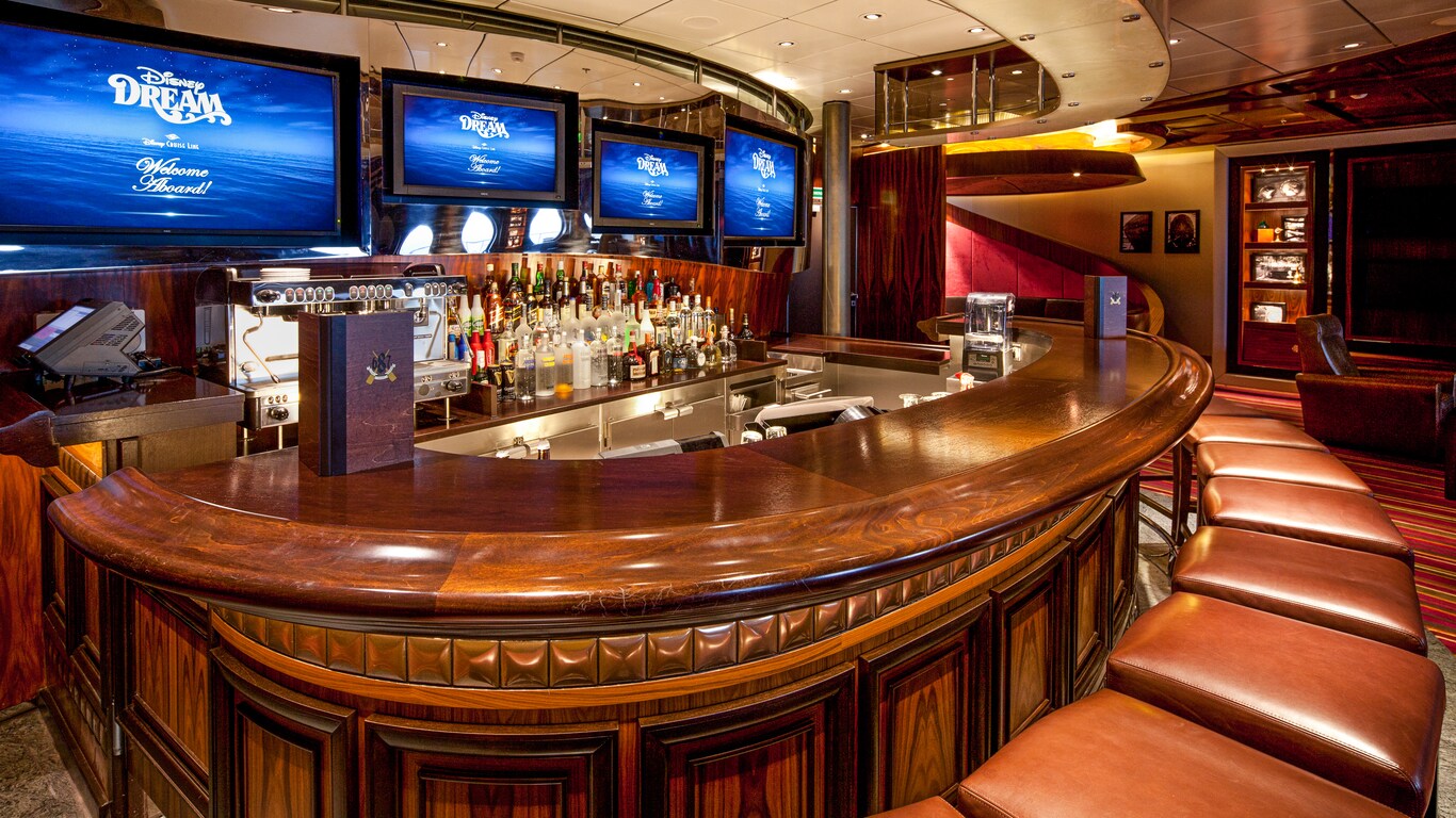 A fully stocked bar in a semi circle shape, with flat panel TVs and leather barstools