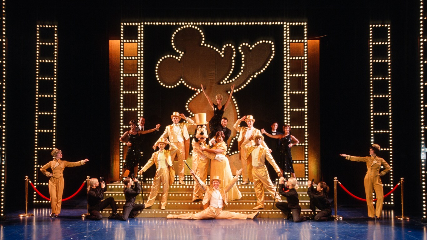 Dancers in sparkling tuxedos and top hats pose on stage with Goofy at The Golden Mickeys