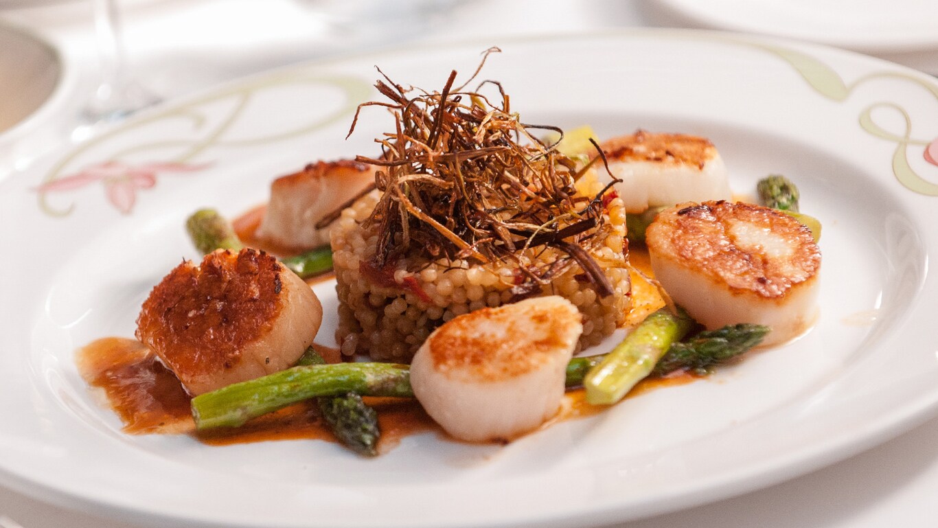 A dish with seared scallops and asparagus surrounding a risotto tower topped with crispy onion