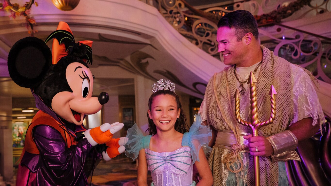 Minnie Mouse in a Halloween costume greeting a young girl and her dad aboard a Disney ship