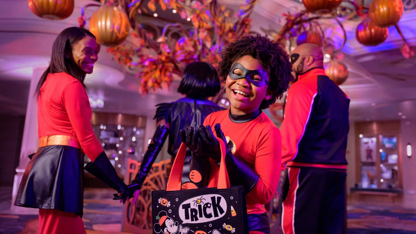 A young boy dressed as a Super Hero holds a trick or treat bag while standing in the atrium of a Disney ship with his family