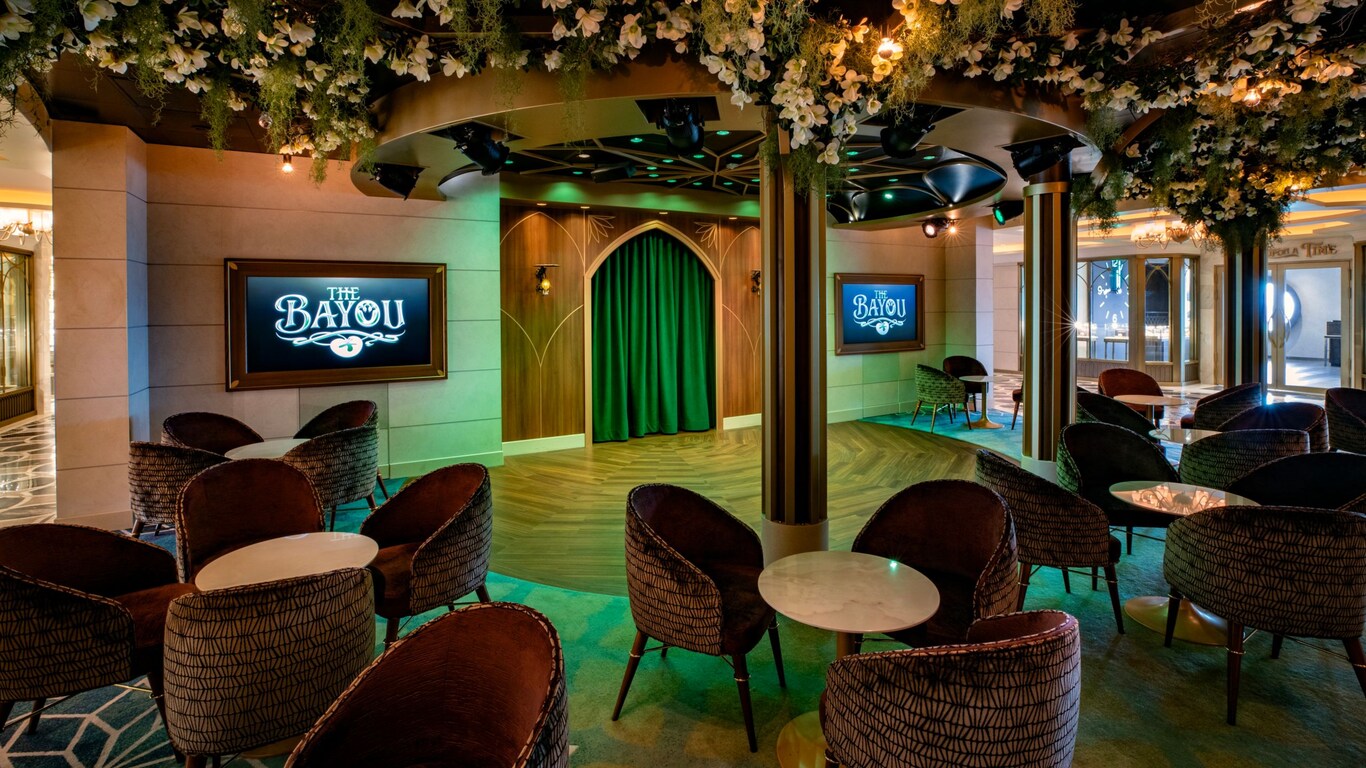A lounge with a seating area around a central stage with screens reading ‘The Bayou’ and a ceiling adorned with artificial foliage and flowers