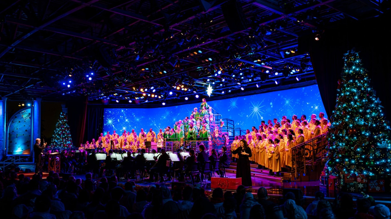 A conductor leading a large choir and orchestra on a festive holiday stage flanked by decorated Christmas trees