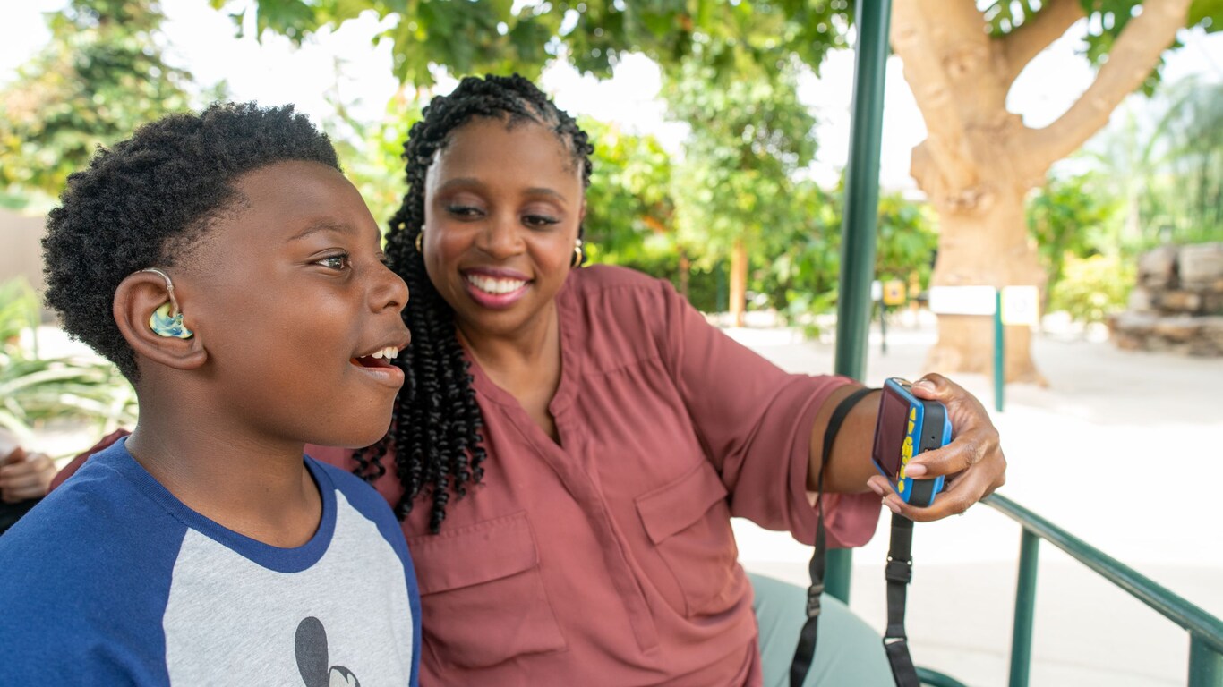 A boy with a hearing aid looks in awe while a woman holds the Disney Handheld Device in front of him.