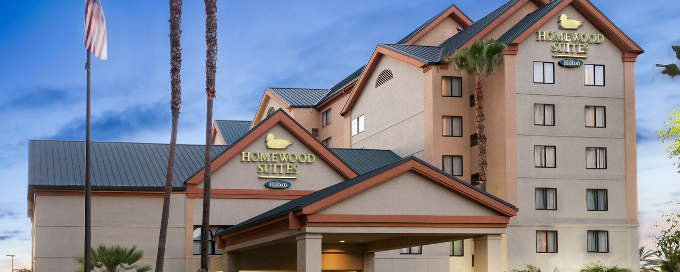 An outside view of Homewood Suites Hilton with landscaping that includes palm trees and an American flag. 