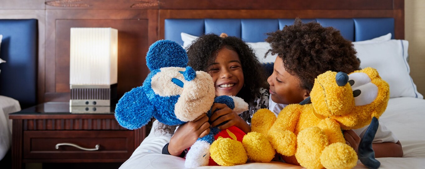 Two children smile and cuddle Mickey Mouse and Pluto plush toys while relaxing on a hotel bed at the Disneyland Hotel