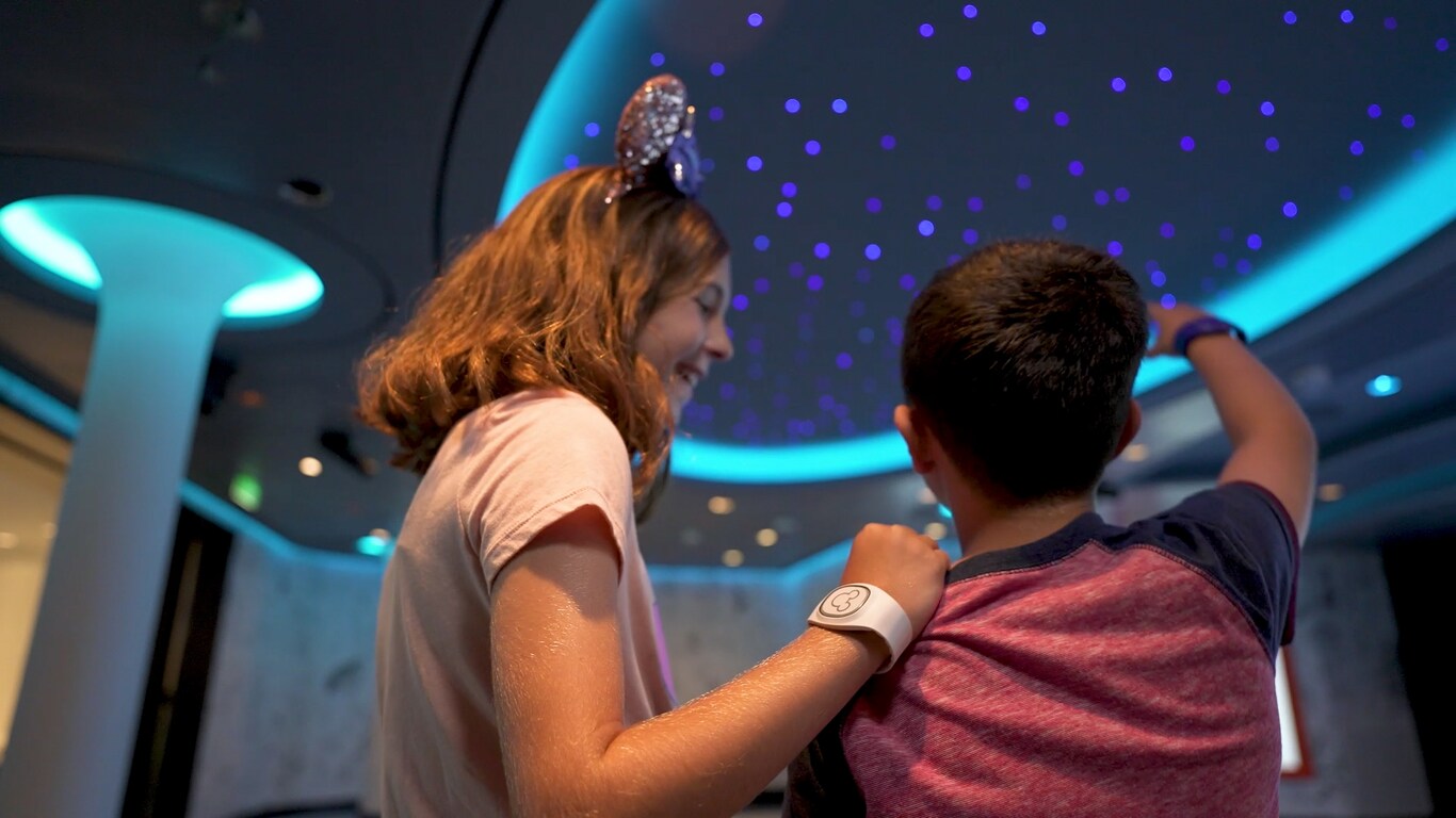 A girl wearing a DisneyBand plus stands next to a boy, pointing at lights projected on the ceiling