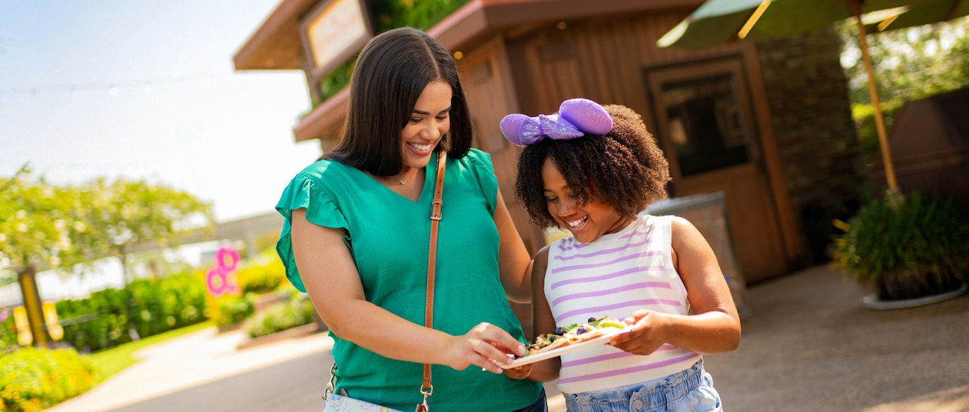 A smiling woman and a girl in a Minnie Mouse ear headband share food outdoors