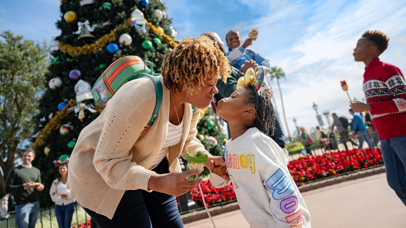 A mother bending down to kiss her daughter in front of a Christmas tree
